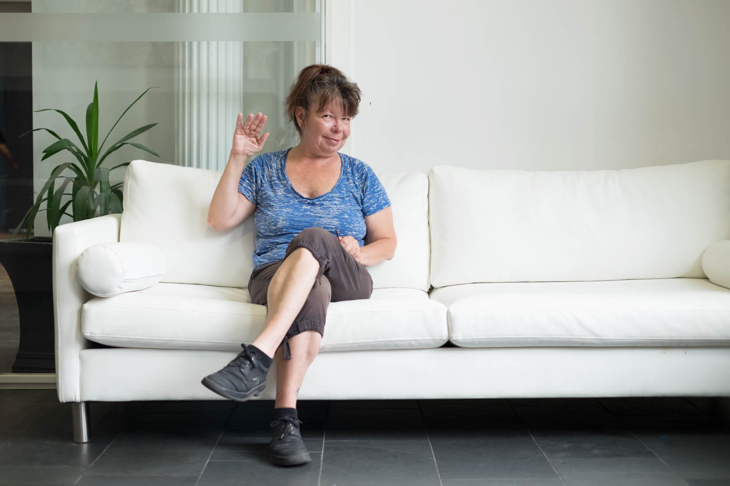 Colleen wearing a blue T-shirt, brown capris, and black laced shoes sitting on a white couch and waving to the camera. Colleen has their hair in a pony tail and there is a white pillar and a potted plant in the background.