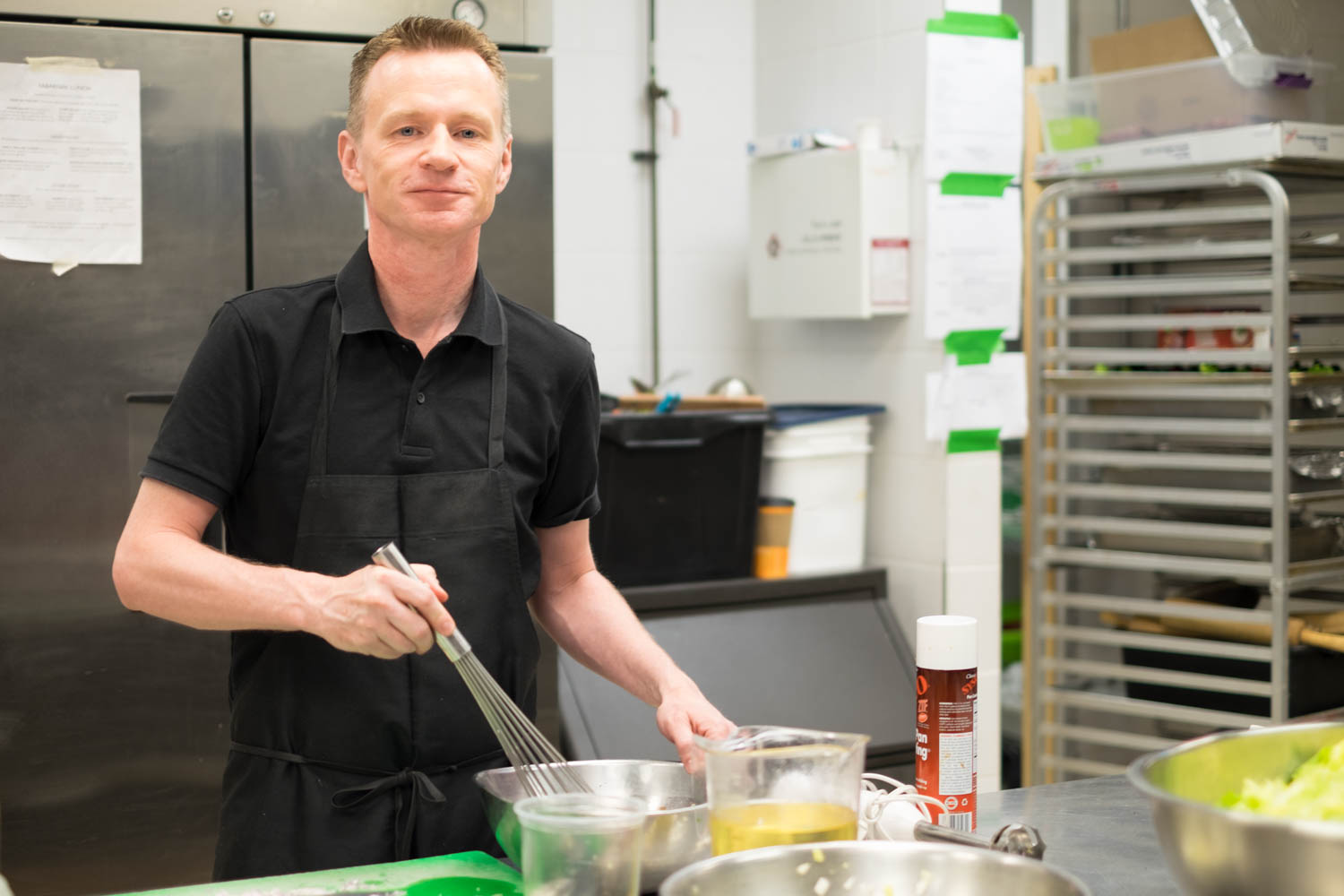 Stephen wearing a black shirt and apron whisking in a kitchen with kitchen equipment in the background. Stephen is looking at the camera and smiling.
